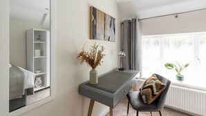 Een moderne slaapkamer met een bed, bureau, stoel, spiegel, plank, schilderij, lamp en raam met een plant.