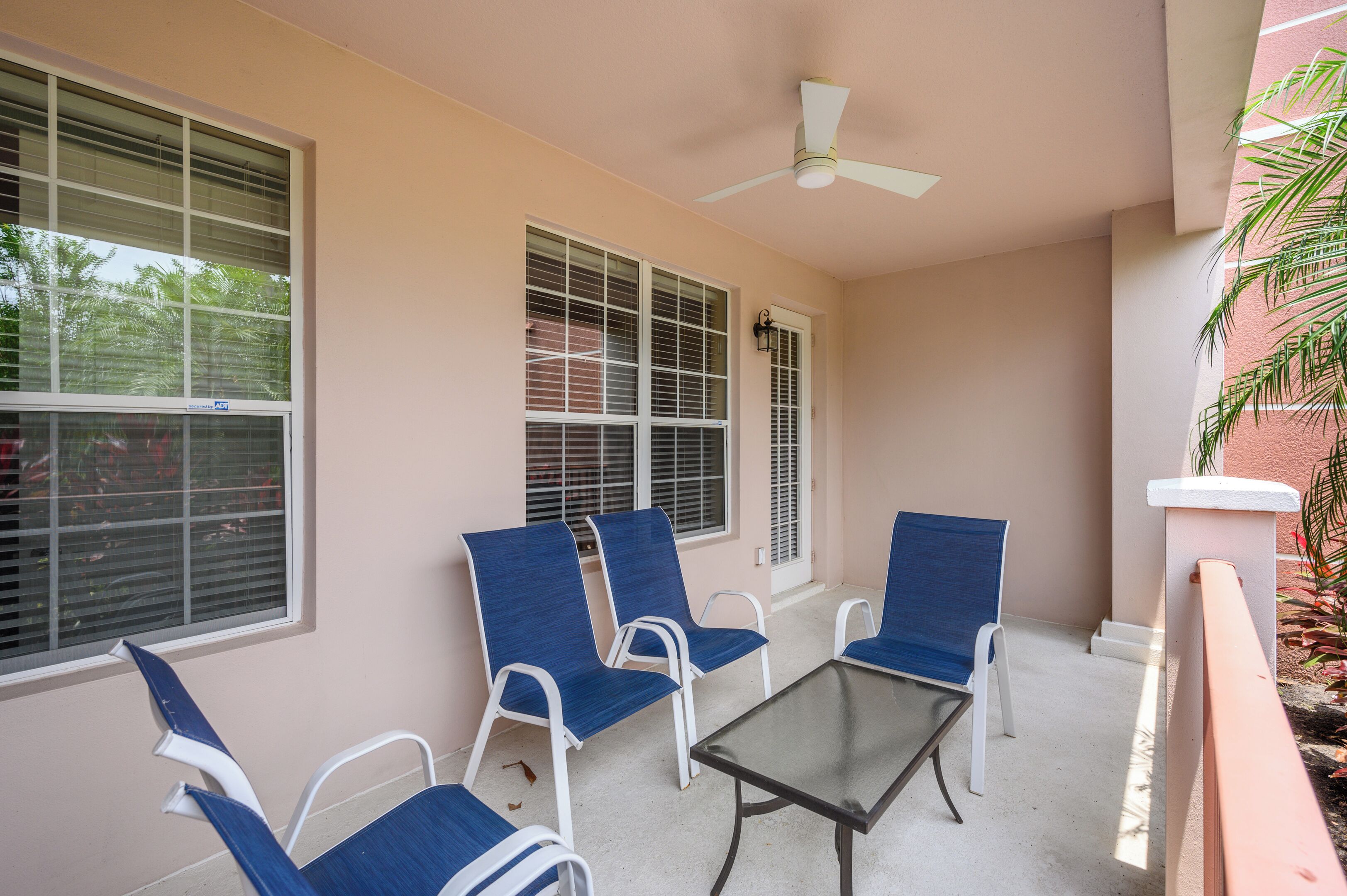 A covered outdoor seating area with four blue chairs and a glass table, large windows with blinds, and a ceiling fan.