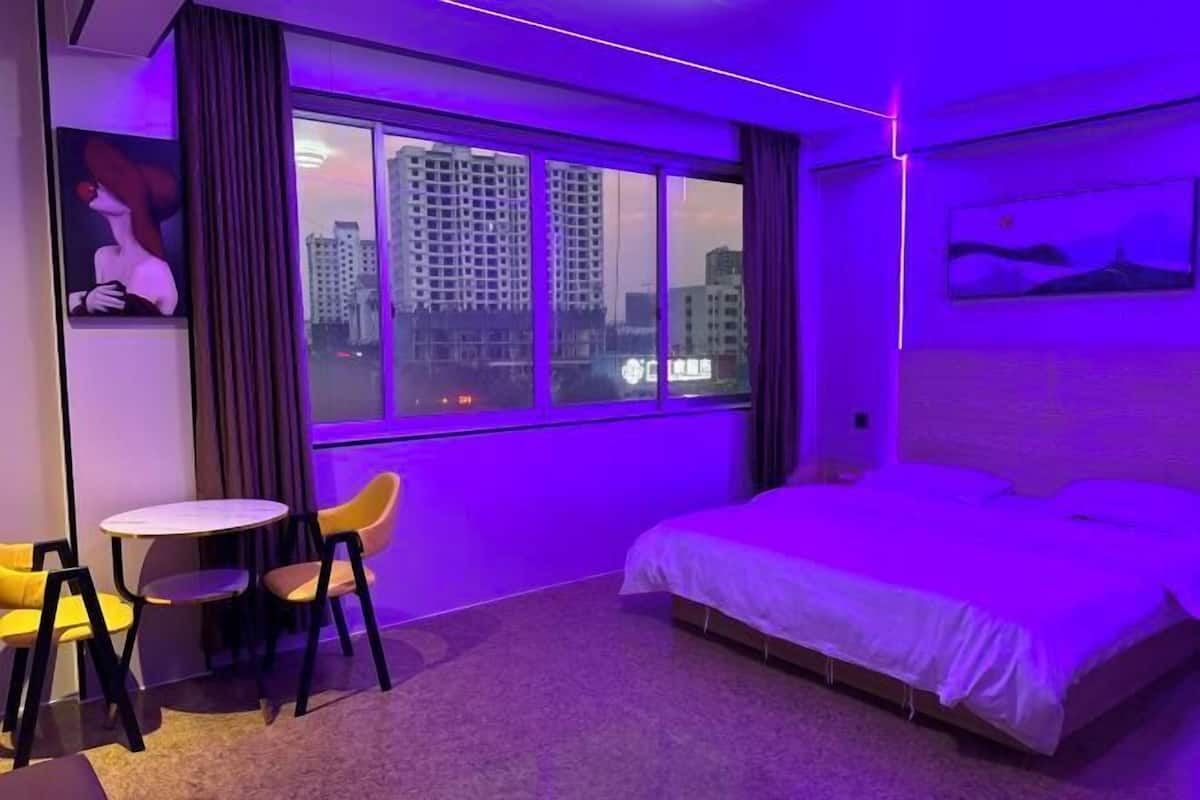 A hotel room with a large window, a bed, a small table with chairs, and a painting on the wall.