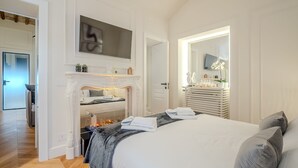 Et moderne soveværelse med pejs, seng med hvid og grå sengetøj, og et stort spejl over kommoden.