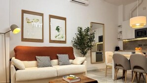 Una sala de estar moderna con un sofá, una mesa de centro y sillas. Se observa una planta, una obra de arte en la pared y una cocina al fondo.