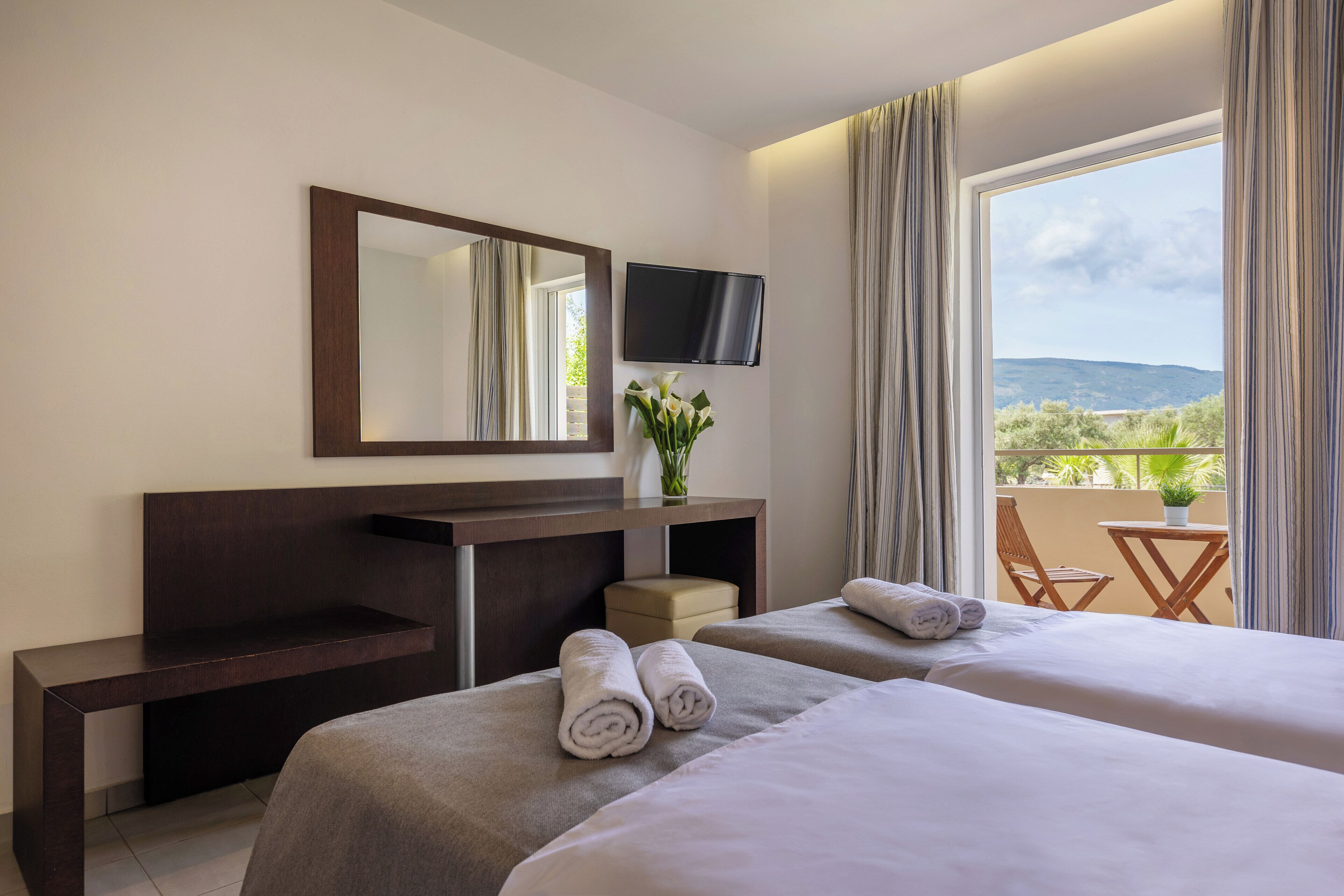Une chambre d’hôtel avec deux lits, une télévision, un bureau et un balcon équipé d’une table et d’une chaise.