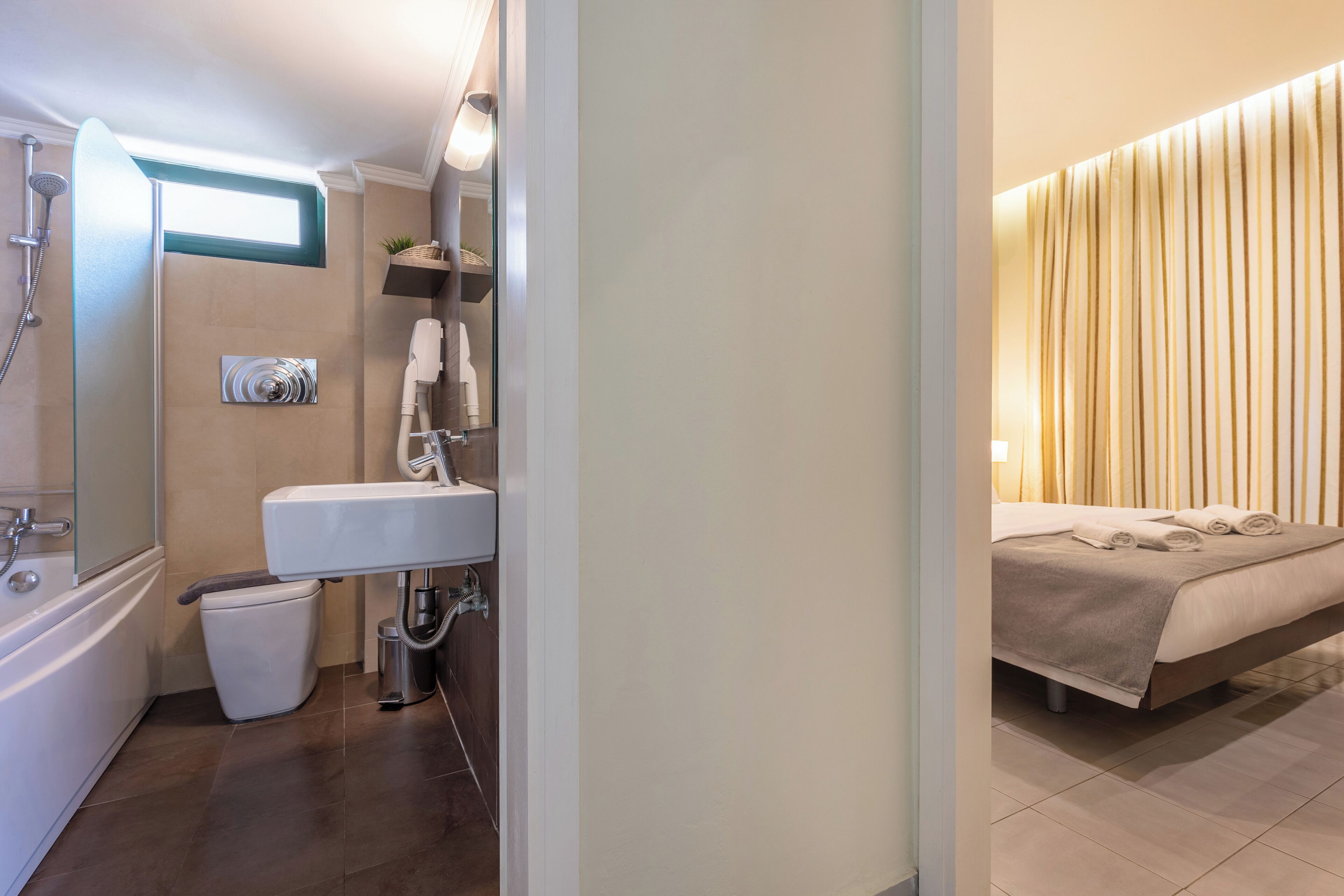 Un baño moderno con ducha, lavamanos e inodoro. Hay una cama con cubrecama color beige y un estante en la pared con toallas.