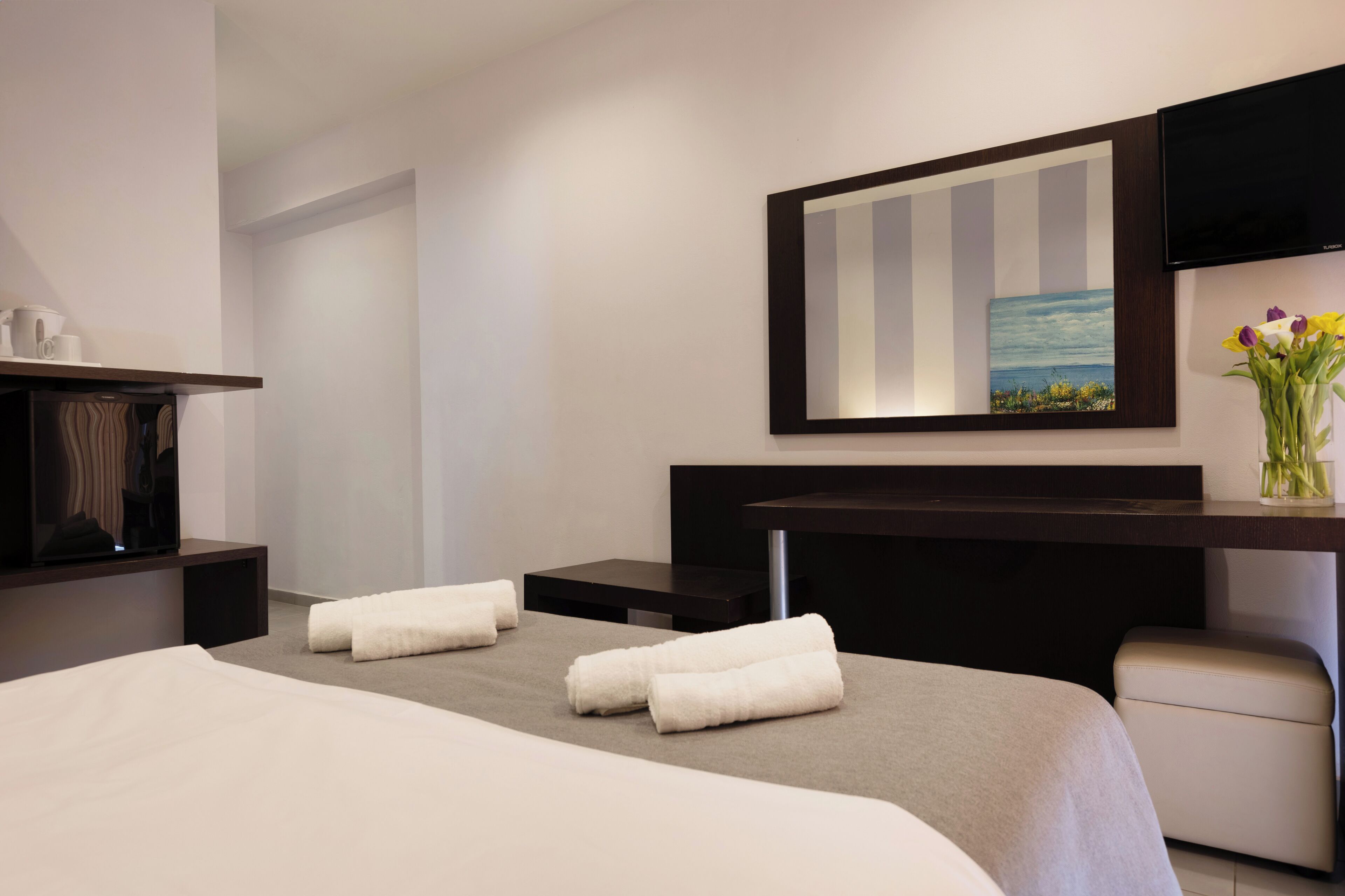A hotel room with a bed, a desk with a flat-screen TV, a painting on the wall, and a vase with flowers.