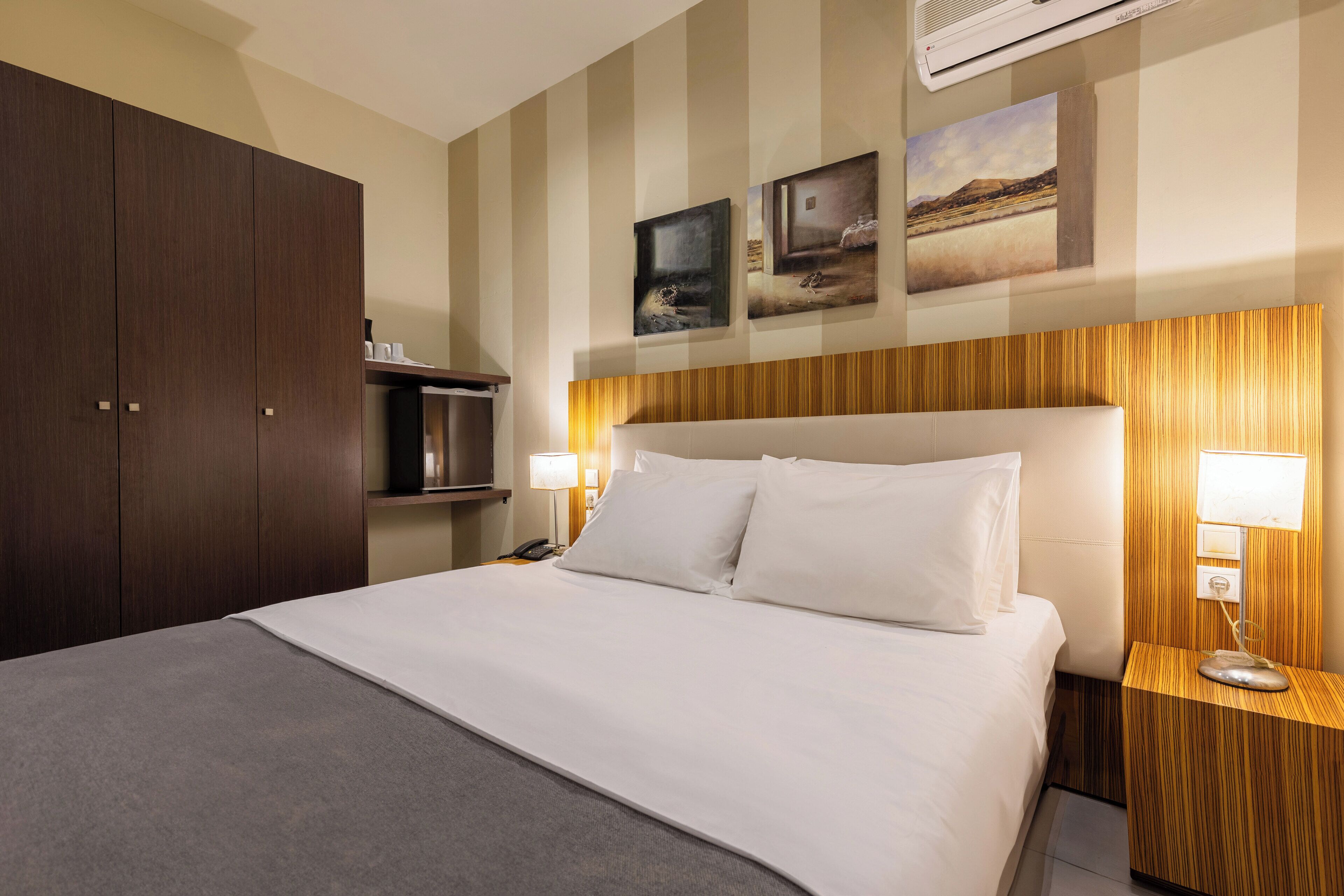 A hotel room with a large bed, wooden headboard, two wall-mounted paintings, a TV, and a minibar.