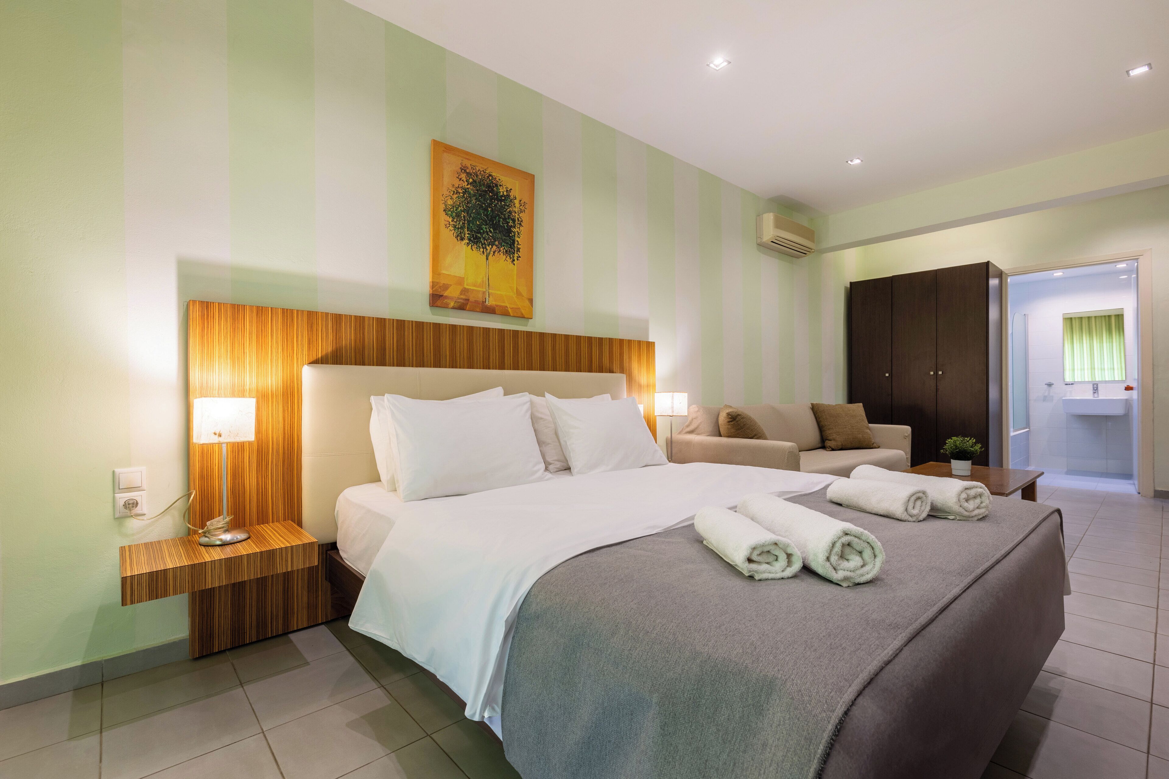 A hotel room with a large bed, a wooden headboard, a bedside table with a lamp, a sofa, and a painting on the wall.