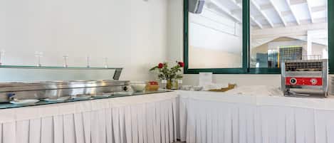 A buffet setup with stainless steel serving dishes, a coffee machine, and a large window with a view of the roof structure.