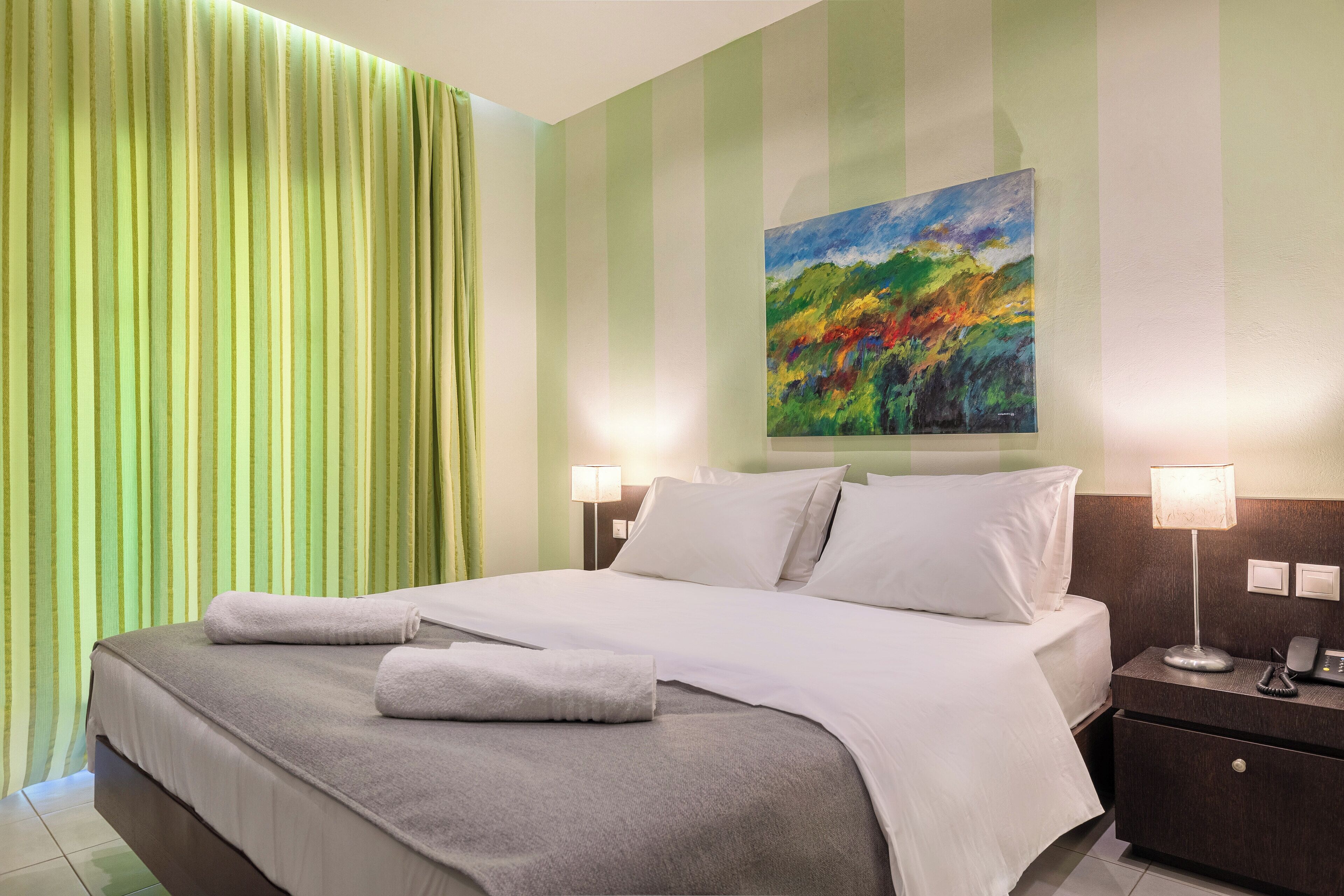 A hotel room with a bed, two bedside lamps, a painting on the wall, and a green curtain.
