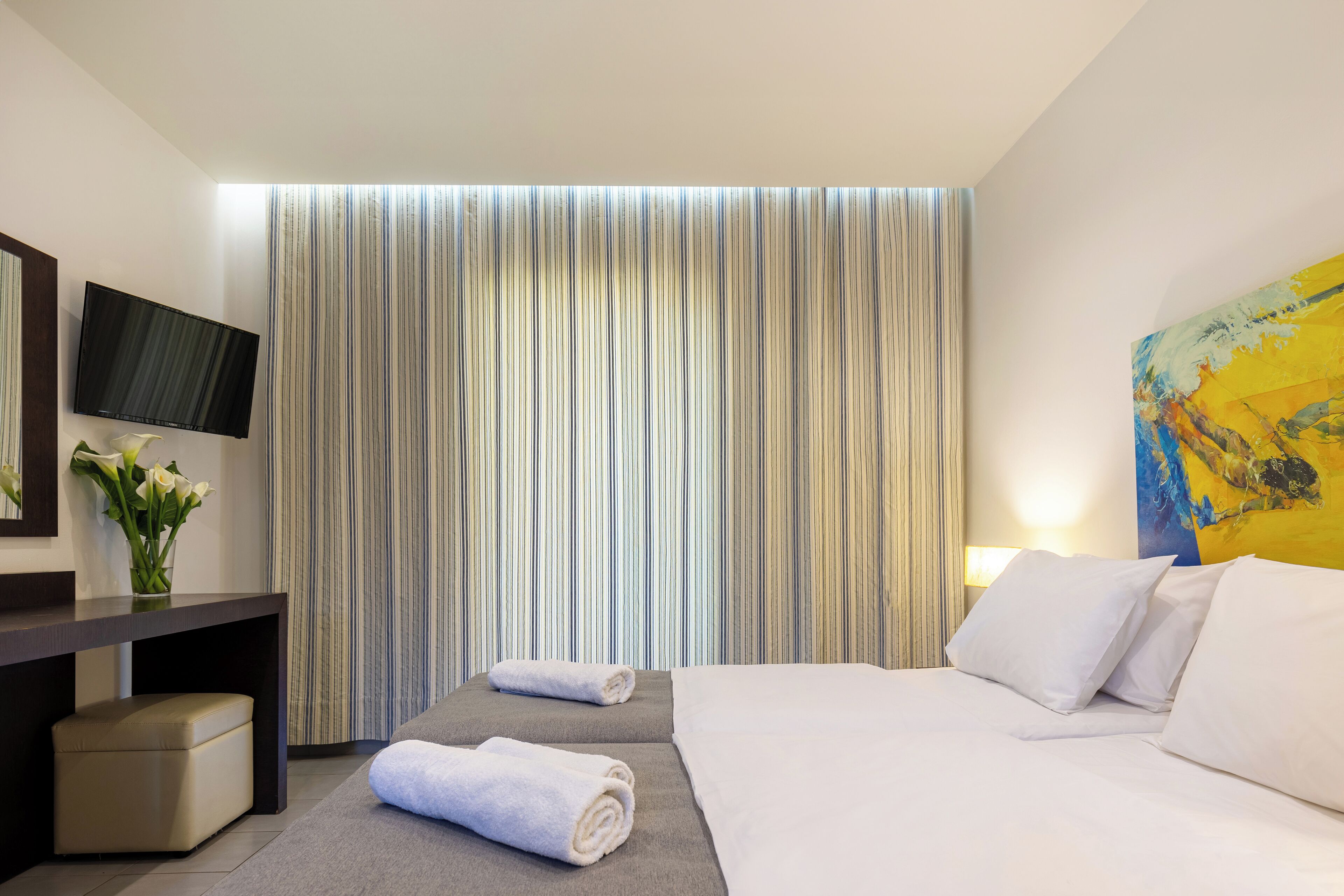 A hotel room with a bed, a desk with a flat-screen TV, a vase with flowers, and a painting on the wall.