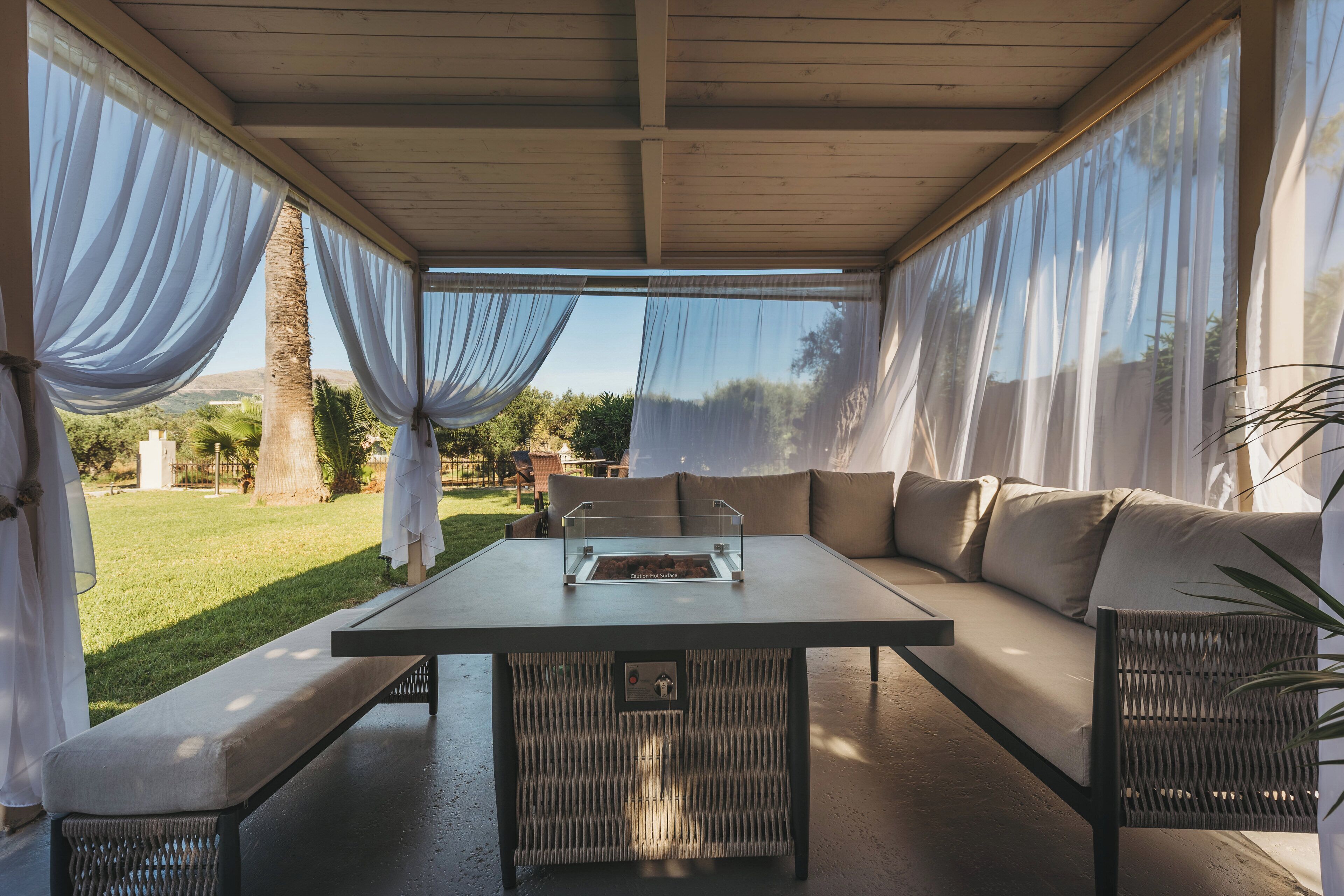 A covered outdoor seating area with a table and chairs, a cushioned sofa, and a view of the surrounding landscape.