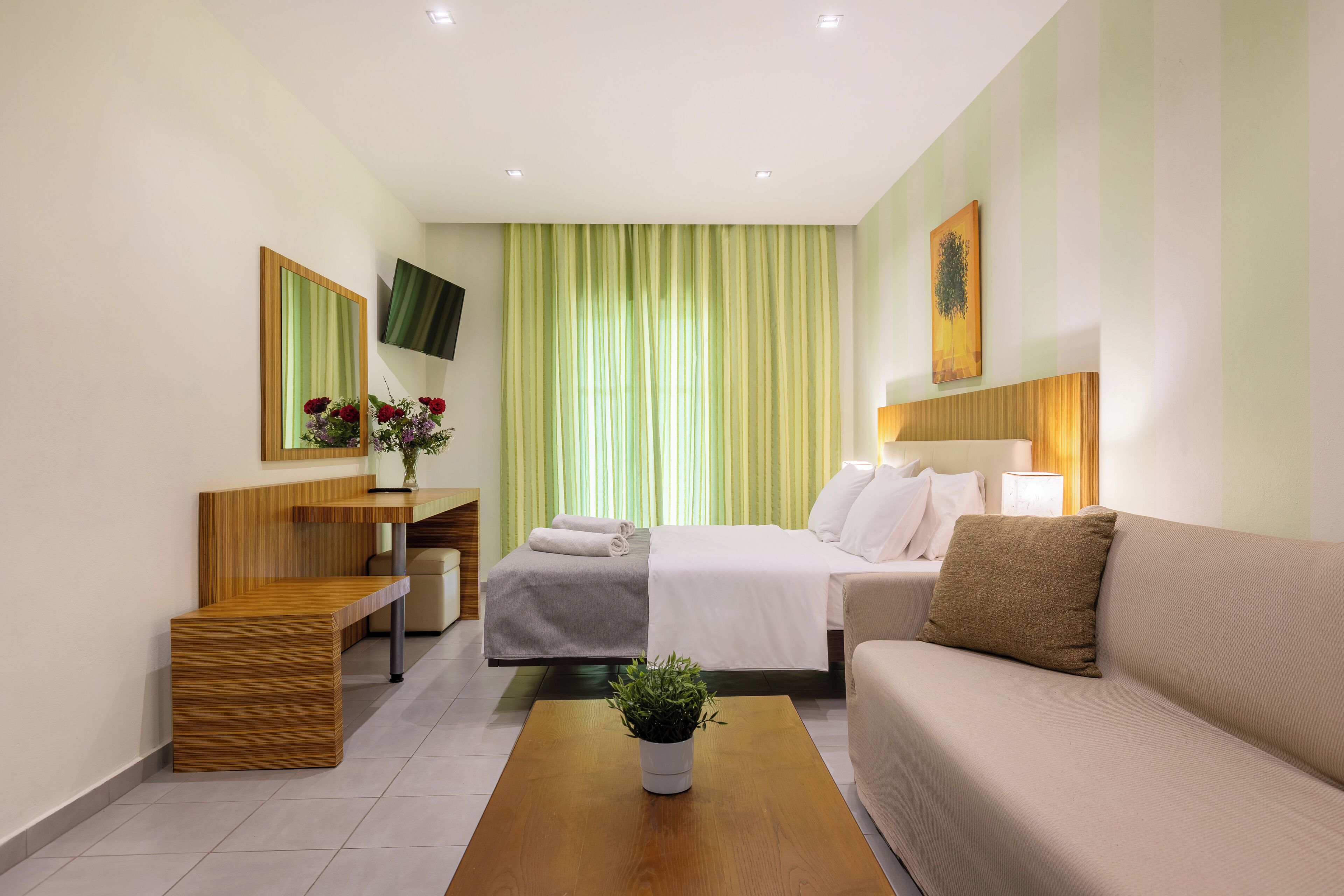 A hotel room with a bed, a sofa, a desk with a mirror, a TV, and a potted plant.