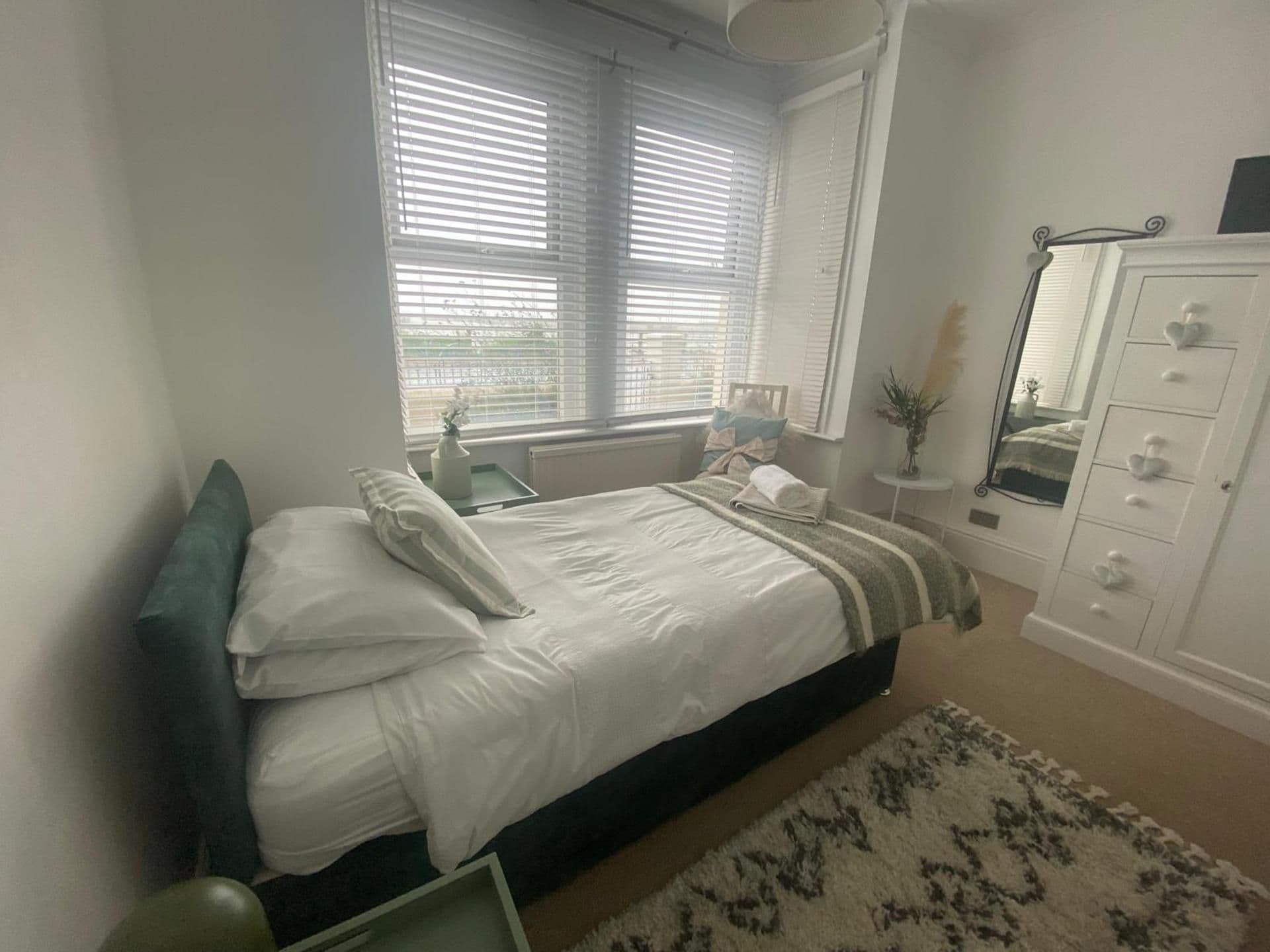 A bedroom with a bed, bedside table, dresser, and a window with blinds.