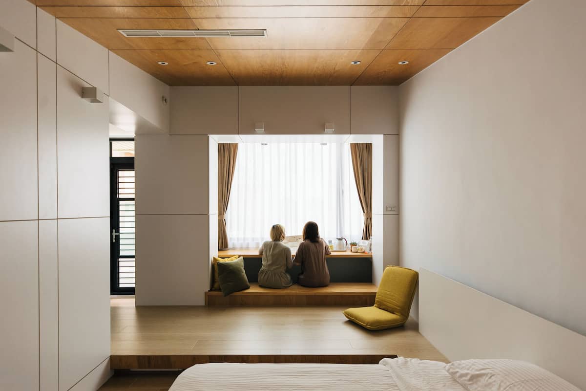A modern bedroom with a wooden ceiling, a large window with curtains, a yellow chair, and a bed with white bedding.