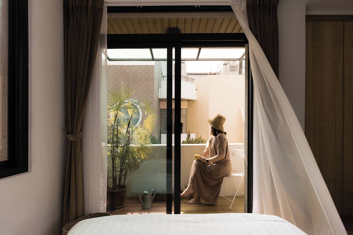 A room with a bed, a window with curtains, a potted plant, and a chair outside.