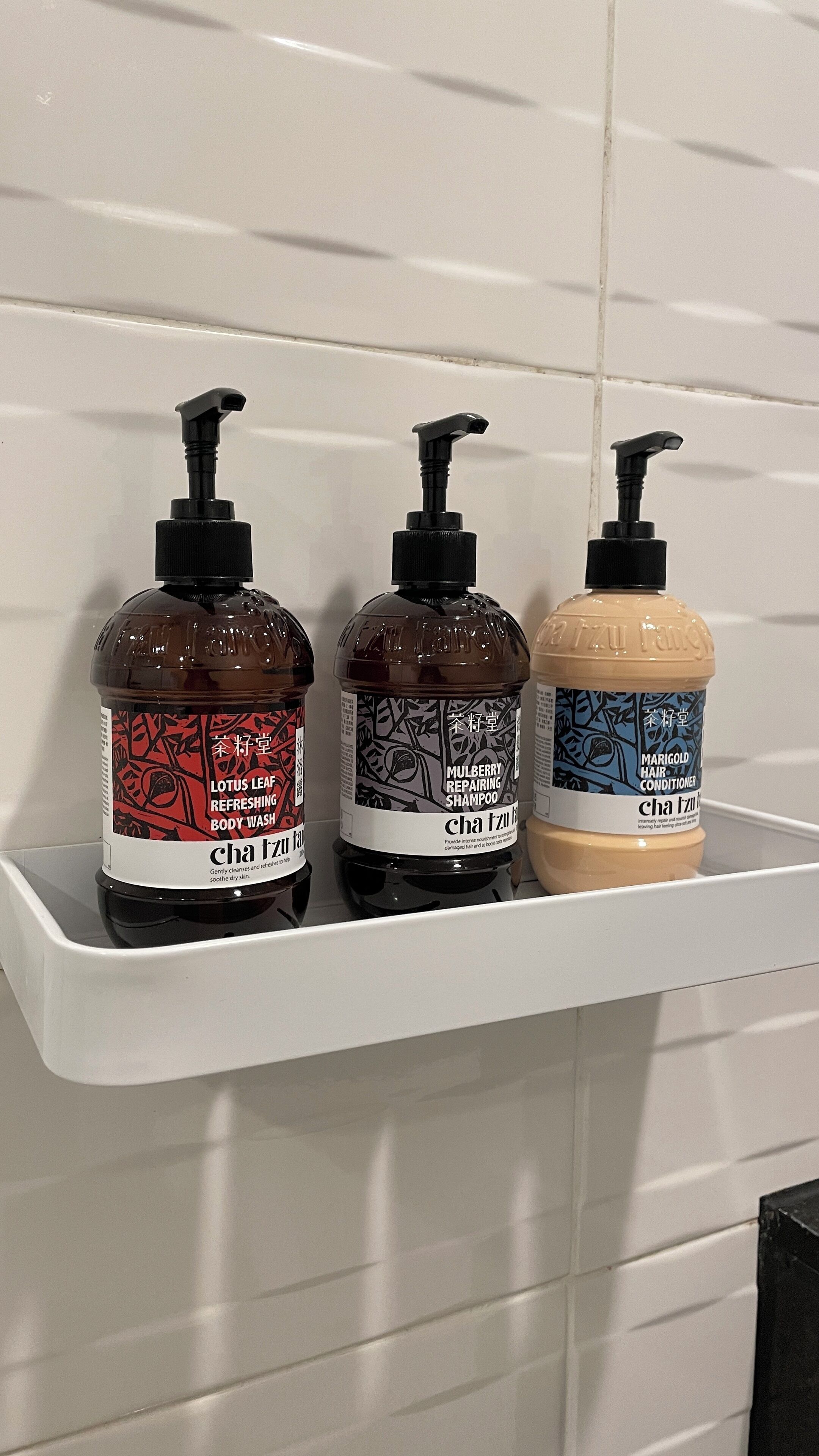 Three bottles of body wash on a white shelf against a tiled wall.