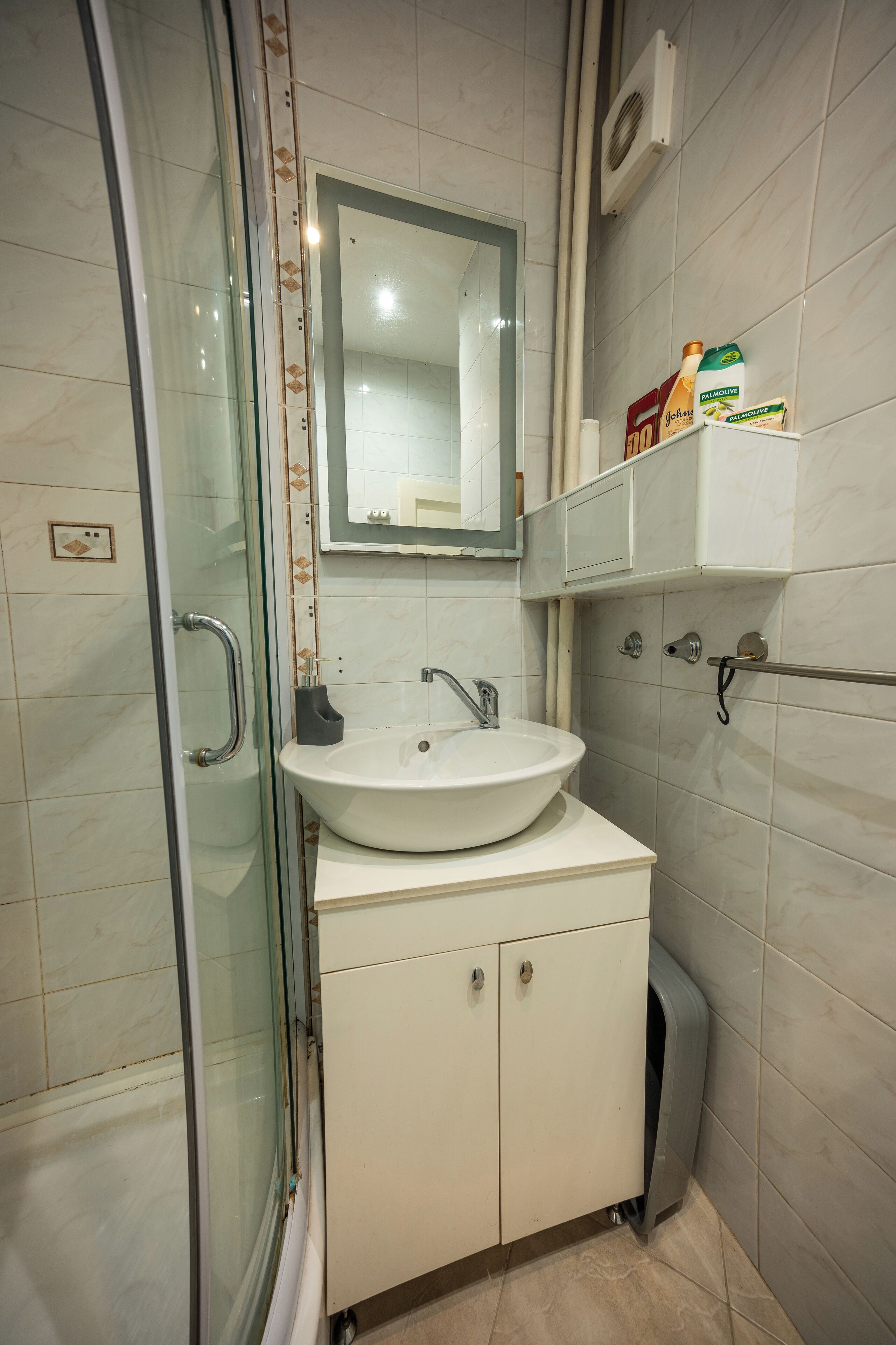 A bathroom with a glass shower enclosure, a white sink, and a mirror above it. There are toiletries on a shelf above the sink.