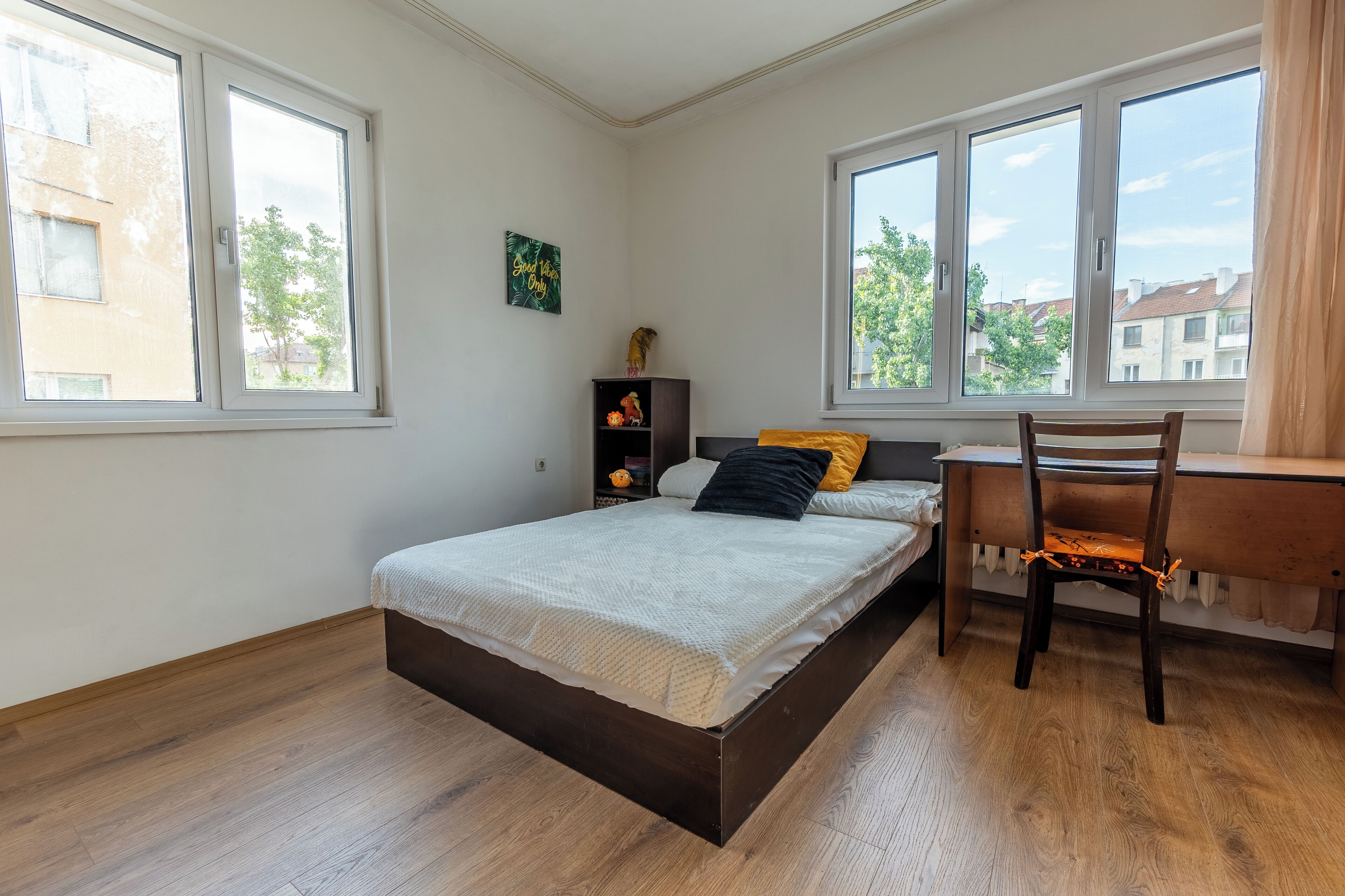A bedroom with a bed, a desk, and a chair. There are windows and a view of buildings outside.
