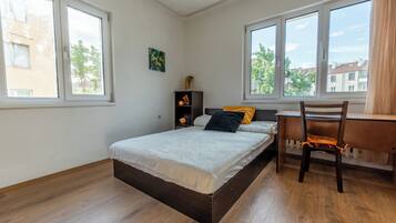 A bedroom with a bed, a desk, and a chair. There are windows and a view of buildings outside.