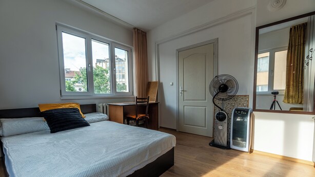 A bedroom with a bed, a desk, a standing fan, and a window with curtains.