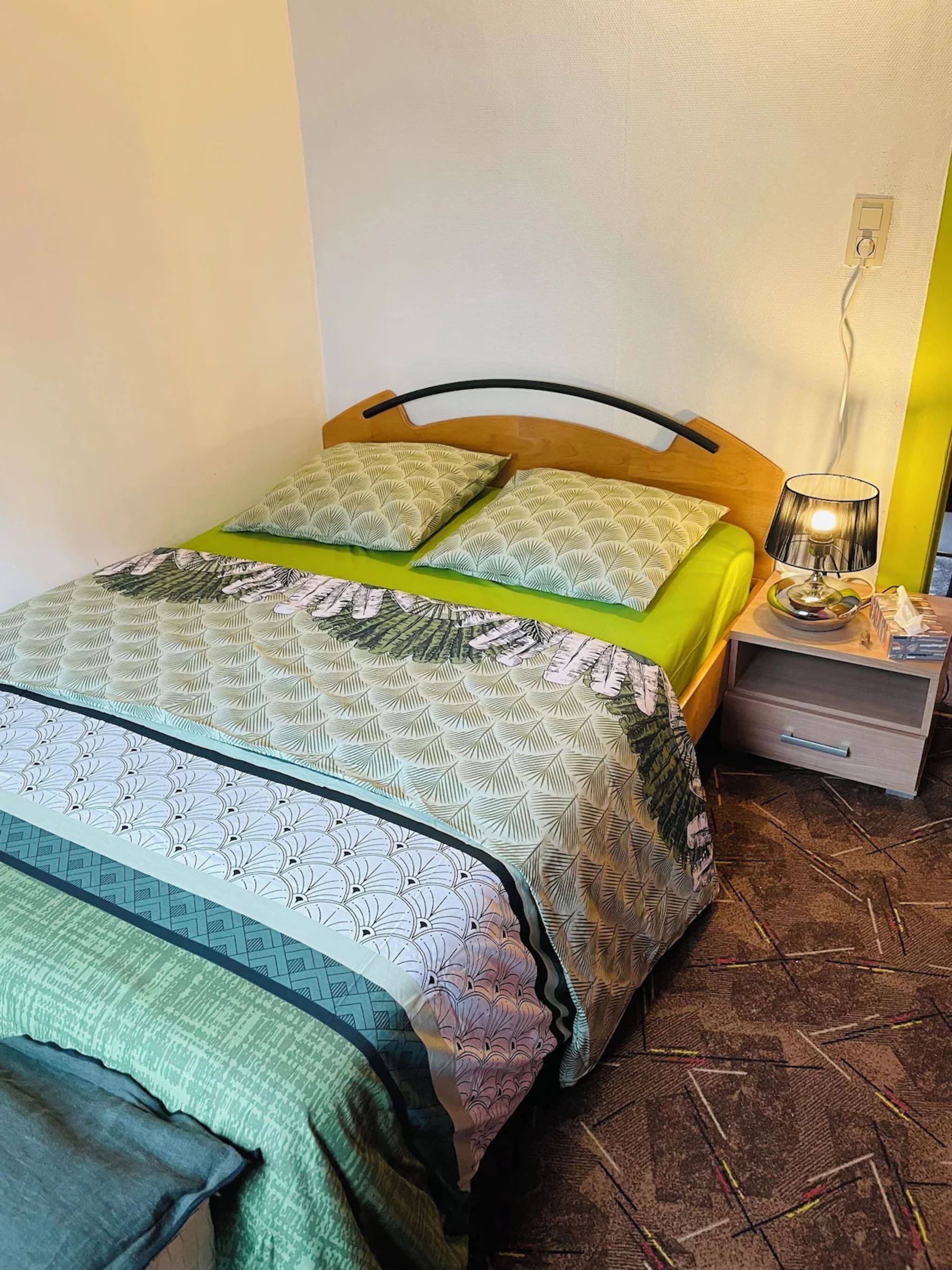 Una cama con ropa de cama estampada, una mesita de noche con lámpara y un enchufe en la pared.
