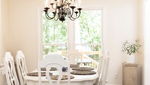 A dining room with a white table, six chairs, a chandelier, and a view through a large window. - Spacious Woodbridge Cottage (Evans)