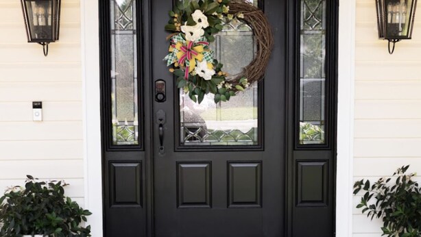 A black front door with a decorative wreath, two wall-mounted lanterns, and potted plants on either side. - Spacious Woodbridge Cottage (Evans)