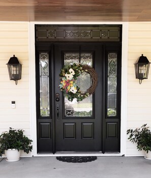 A black front door with a decorative wreath, two wall-mounted lanterns, and potted plants on either side. - Spacious Woodbridge Cottage (Evans)