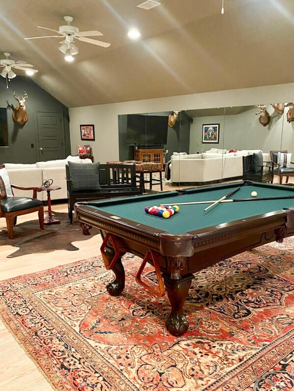 A pool table in a room with a patterned carpet, a sofa, and a television. - Elegant Stonegate Home (Evans)