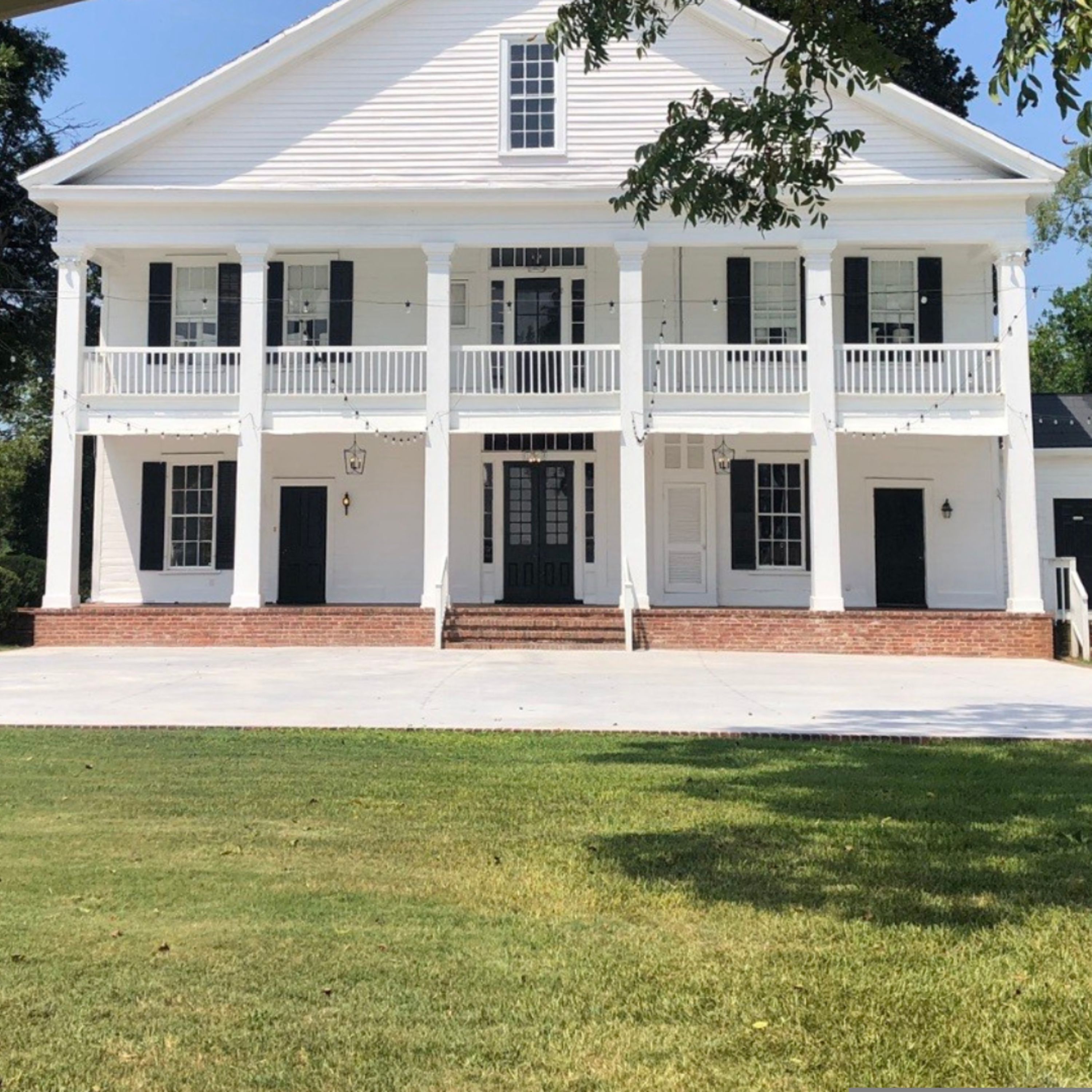 A two-story white building with a central entrance, a balcony on the upper floor, and a brick foundation.