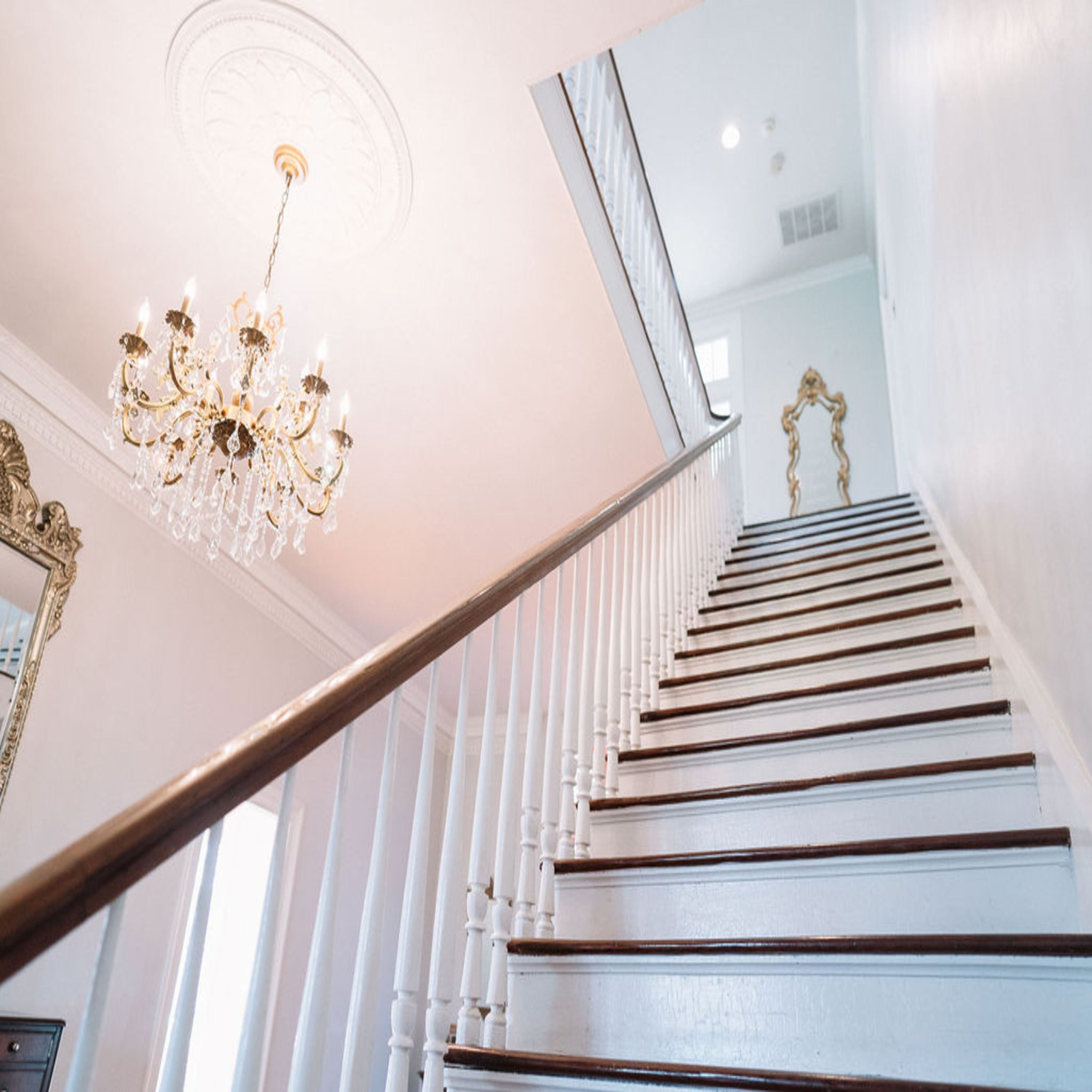 A staircase with white balusters and a wooden handrail, leading to a room with a chandelier.