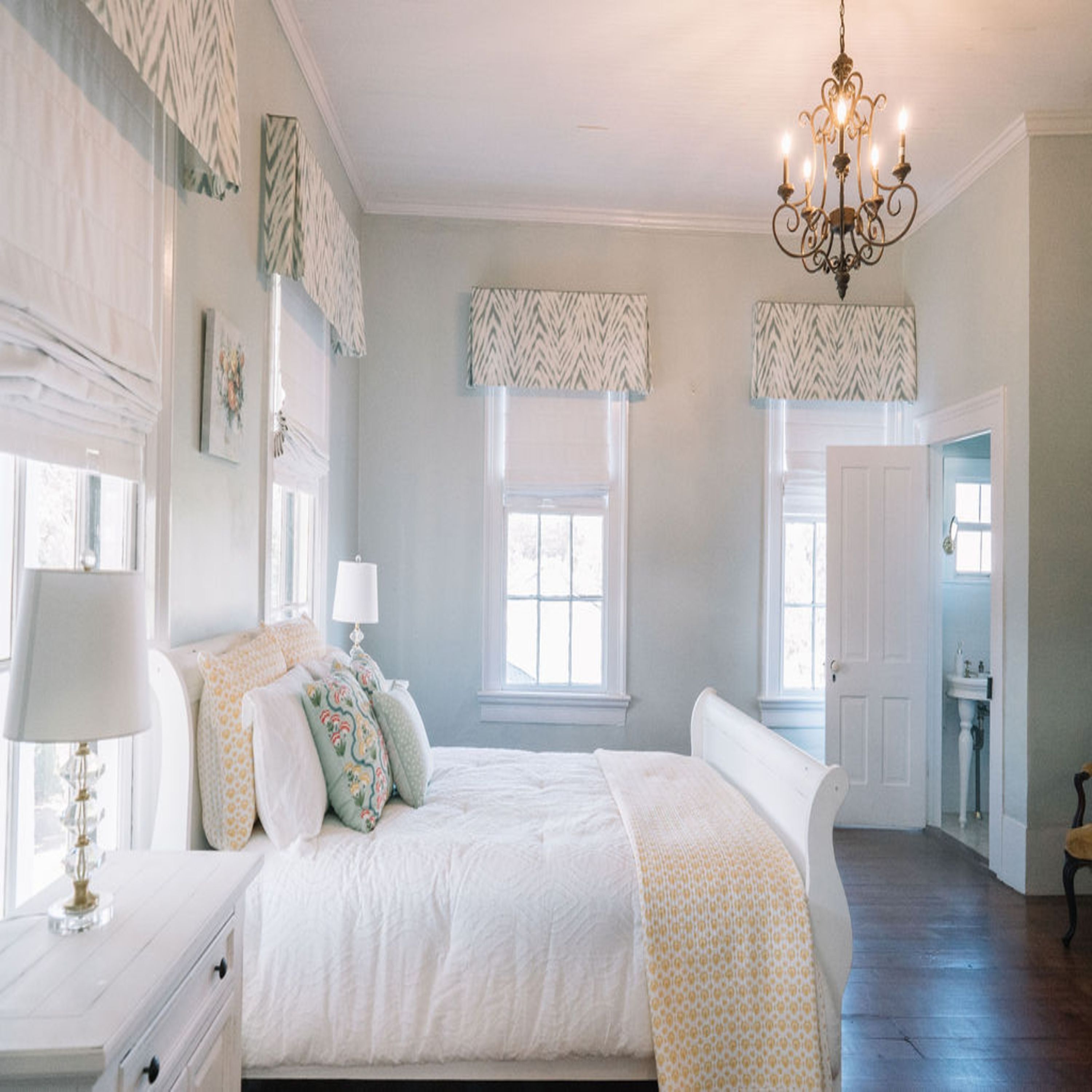 A bedroom with a large bed, a dresser, a nightstand, a chandelier, and two windows with patterned valances.