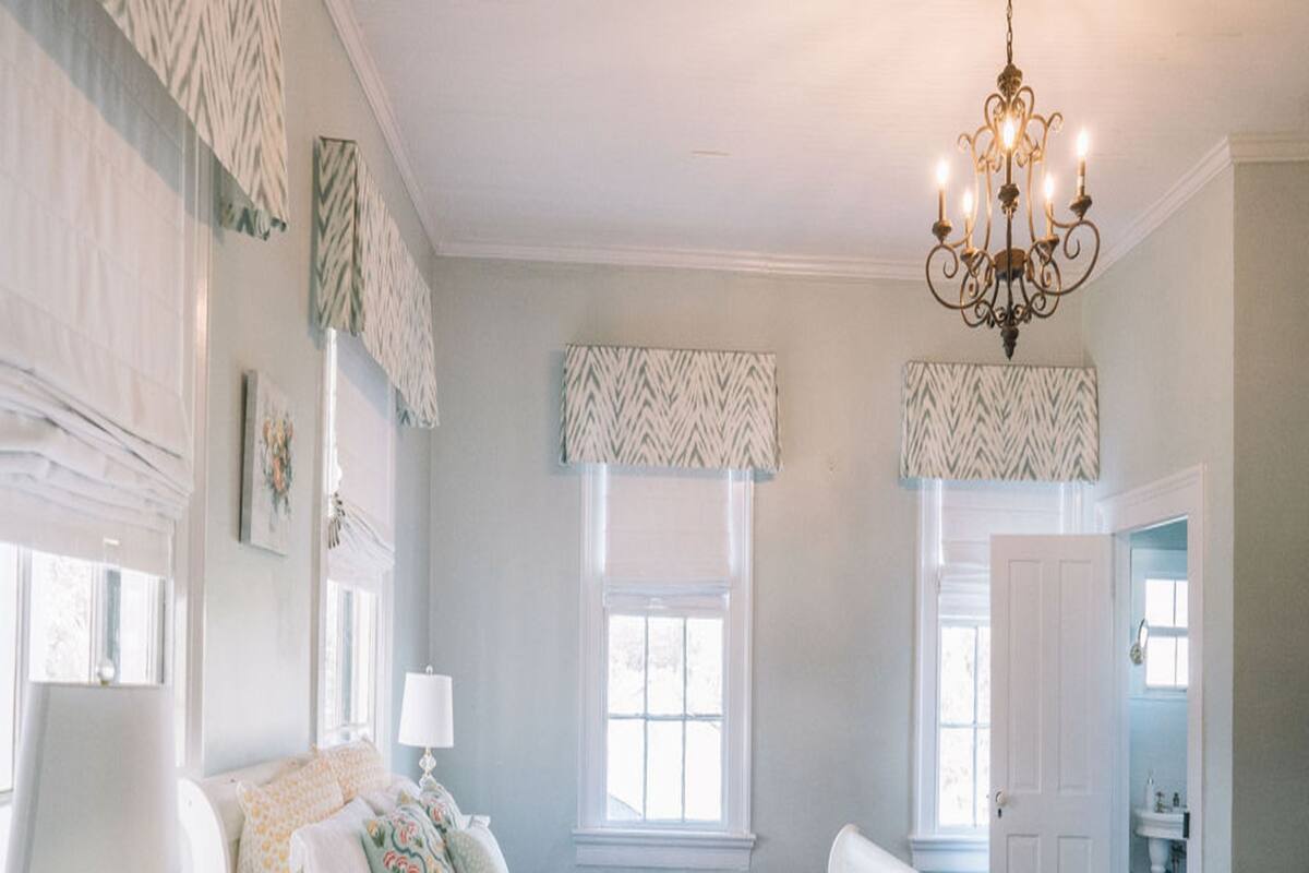 A bedroom with a large bed, a dresser, a nightstand, a chandelier, and two windows with patterned valances.