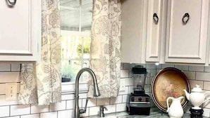 A kitchen with white cabinets, a stainless steel sink, a window with patterned curtains, a blender, and a teapot on the countertop. - Elegant Crofton Ranch (Augusta)