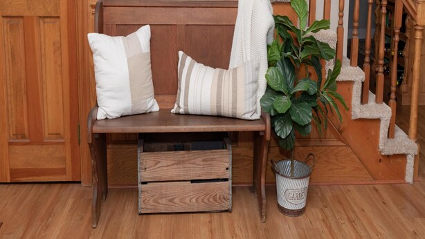 A wooden bench with two pillows, a potted plant, and a metal container labeled "GARDEN" in a room with wooden flooring and a wooden staircase.