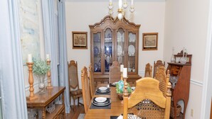 A dining room with a wooden table set for four, surrounded by chairs. A wooden cabinet with glass doors is in the background, and a chandelier hangs above the table.