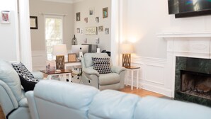 Uma sala de estar com lareira, um sofá, uma poltrona e uma mesa com abajur.