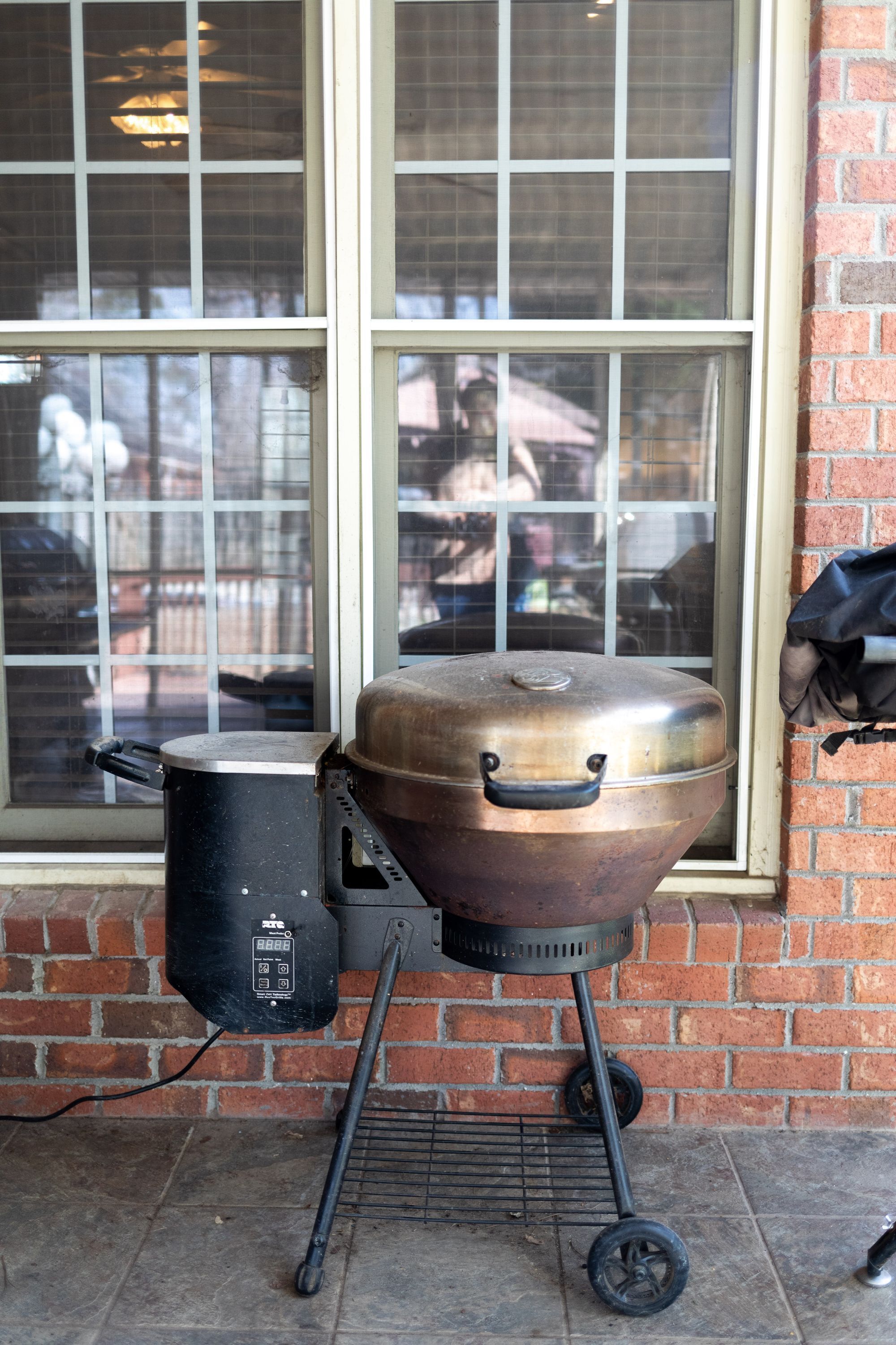 Ein tragbarer Grill mit Deckel und einem schwarzen Topf, vor einer Backsteinmauer und einem Fenster platziert.