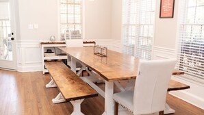 Uma área de refeições com mesa de madeira e bancos, cadeiras brancas e uma janela com persianas.