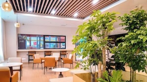 Un espace intérieur moderne avec des plafonds à lattes de bois, de nombreux coins salon comprenant des tables rondes et des canapés, ainsi que de grandes fenêtres ornées d’œuvres d’art colorées.
