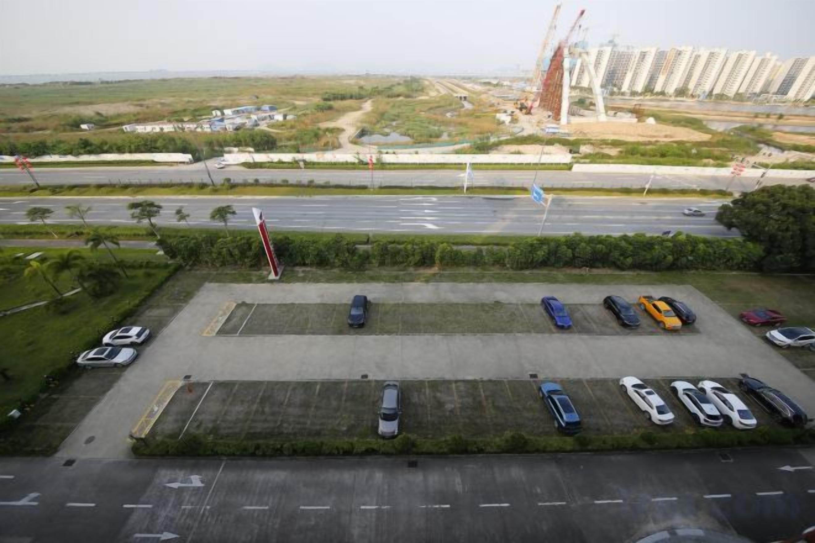 Vista aérea de um estacionamento com vários carros, uma estrada e edifícios ao longe.