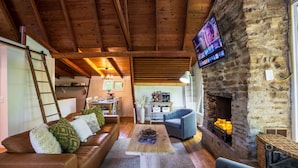 A cozy living room with a stone fireplace, wooden ceiling beams, a television mounted on the wall, and a comfortable seating area with a sofa and armchairs. - Lake View Hot Tub Modern A-frame (Rockbridge)