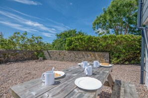 Une table de pique-nique en bois, avec des assiettes, des gobelets et une tasse, est installée à l’extérieur, devant un mur de pierres et sur fond de verdure.