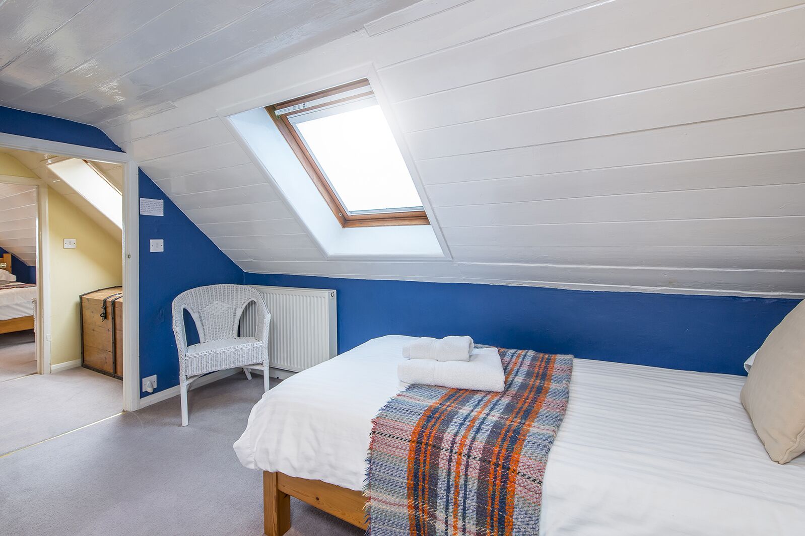 A bedroom with a sloped ceiling, a skylight, a bed with a white duvet and a colorful throw, a white wicker chair, and a wooden chest of drawers.
