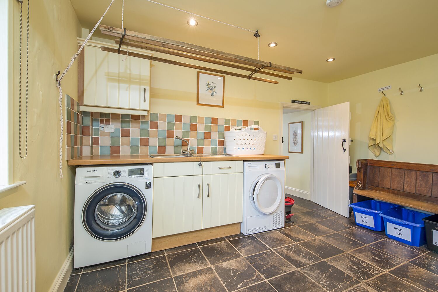 A laundry room with a washing machine, dryer, and a drying rack. There are blue storage bins labeled "Boot Box" and a yellow towel hanging on a hook.
