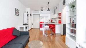 Eine moderne Wohnung mit Wohnbereich, ausgestattet mit einem roten Sofa, einem kleinen runden Tisch und einem Bücherregal. Die Küche verfügt über rote und weiße Schränke, eine zentrale Kochinsel und Barhocker.