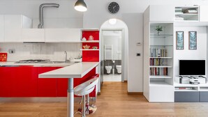Eine moderne Küche mit einer roten Kochinsel, weißen Schränken und einem integrierten Fernseher. Ein Flur führt zu einem Badezimmer und einem Wohnbereich mit Bücherregalen.