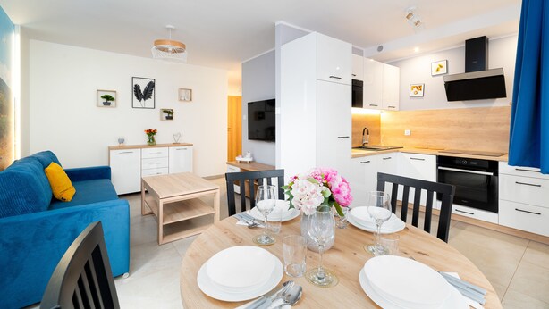 A modern living room with a blue sofa, a dining table set for four, and a kitchen area with white cabinets and a black oven. - Apartamenty Sun & Snow Nad Łomnicą (Karpacz)