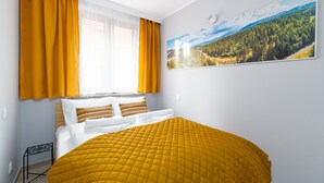 A bedroom with a bed, yellow curtains, a window with a view of a forest, and a painting of a landscape. - Apartamenty Sun & Snow Nad Łomnicą (Karpacz)