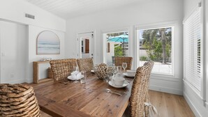 Un coin repas avec une table en bois dressée pour quatre personnes, entourée de chaises en osier, de murs blancs et de grandes fenêtres équipées de stores.