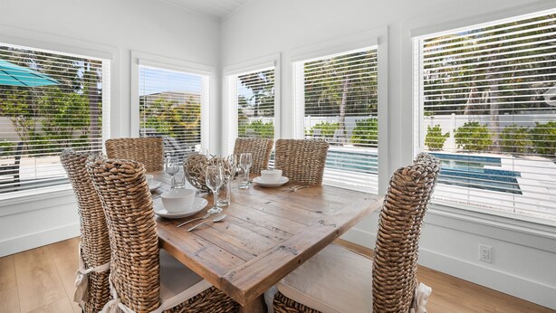 Un coin repas avec une table en bois dressée pour quatre personnes, entourée de chaises en osier, de grandes fenêtres équipées de stores et donnant sur une piscine.